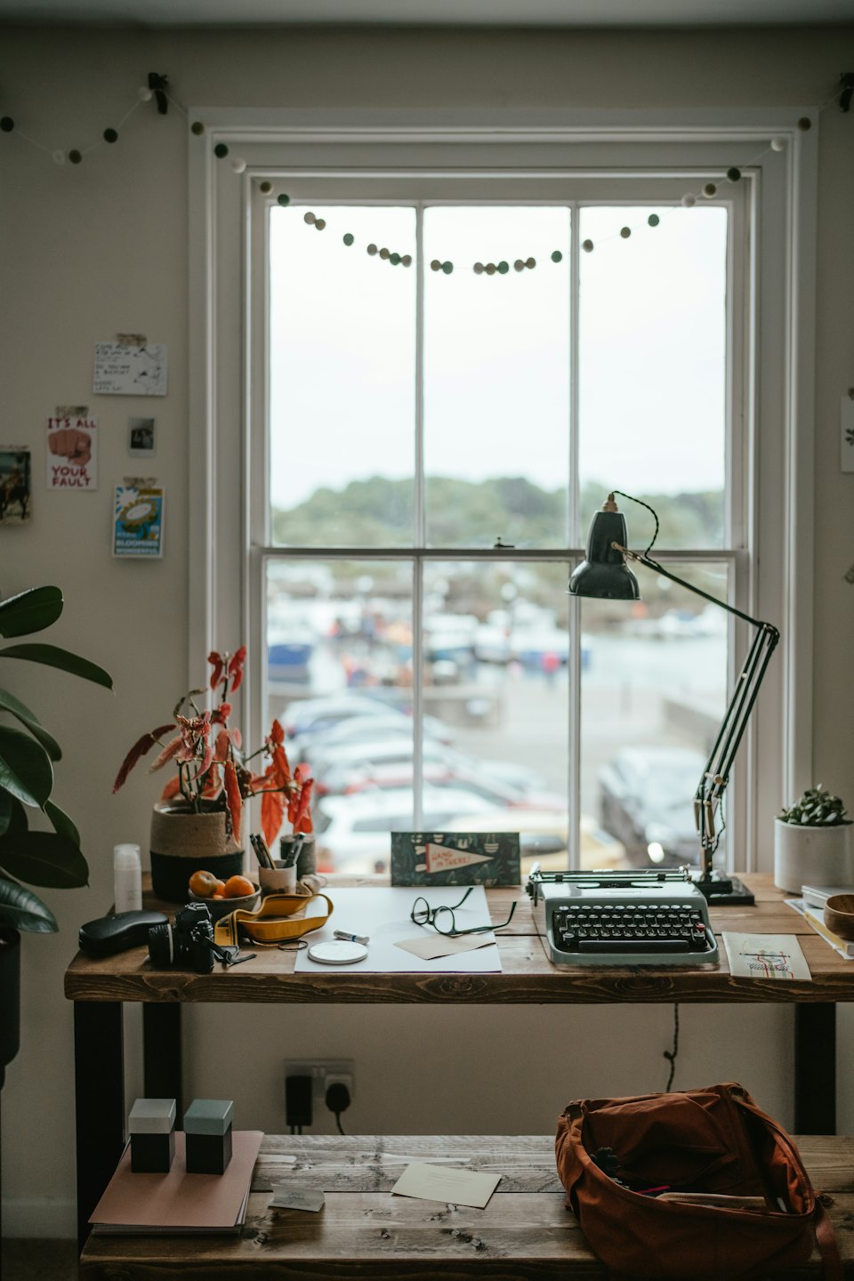 Rustic desk with typewriter and plants near a large window.